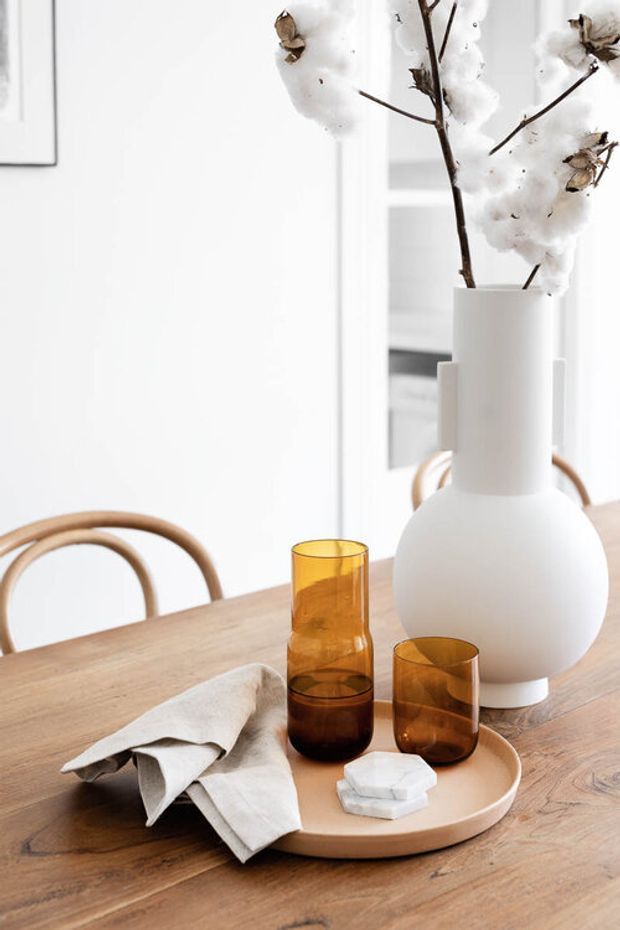 Bright, minimalist dining vignette featuring a light timber dining table, a large matte white sculptural vase with cotton branches, amber glass tumblers, a ceramic serving tray, folded linen napkin, and curved timber dining chairs. White walls and cabinetry create an airy neutral backdrop.