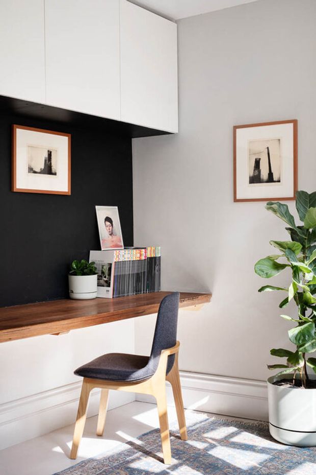 A compact, neatly styled home office nook with a wall-mounted wooden desk, white overhead cabinetry, and a black feature wall. A mid-century-inspired upholstered wooden chair sits at the desk, alongside framed monochrome artwork, magazines, a small plant, and a tall potted fiddle-leaf fig. White lower wall panelling and a patterned rug complete the bright, refined workspace.