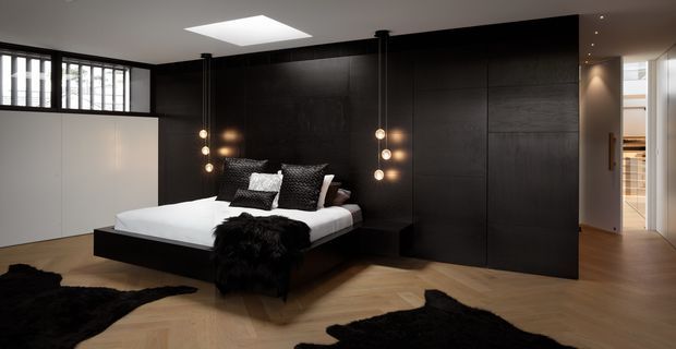 Contemporary master bedroom with a low-profile black bed, crisp white bedding, black textured cushions and throws, and dark full-height built-in wall panelling and cabinetry. Warm herringbone timber flooring contrasts with the dark joinery, while globe pendant lights and a large skylight provide focused illumination. A white built-in wardrobe and hallway are visible at the edges of the room.