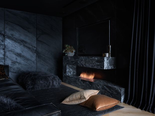 A dark, luxurious bedroom with a black upholstered bed, plush dark bedding, sculptural pillows, dramatic veined stone wall panels, and a matching stone fireplace with a visible flame. Full-height black curtains and low ambient lighting create a moody, intimate atmosphere.