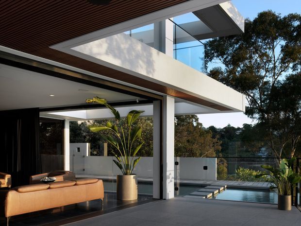 Contemporary two-storey residence with strong indoor–outdoor connection, featuring broad white cantilevered forms, extensive glazing, a slatted timber-lined soffit, and a covered terrace overlooking a narrow swimming pool and landscaped surroundings. The open living area includes a tan leather sofa, dark flooring, large potted plants, and frameless glass pool barriers.