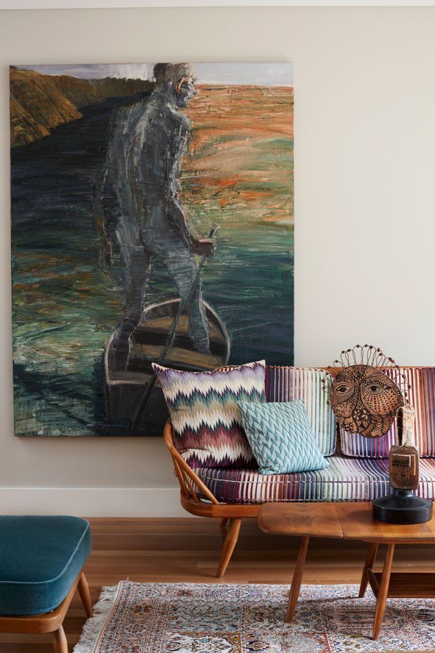 A warmly styled living room featuring a mid-century-inspired upholstered lounge chair or small sofa with striped fabric, patterned cushions, a wooden coffee table, a teal footstool, and a richly patterned rug. A large expressive figurative painting dominates the neutral wall behind the seating, complemented by a decorative woven or carved face artwork. Timber flooring and warm wood furniture create an inviting, eclectic atmosphere.