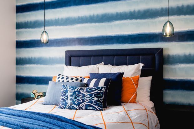 A stylish bedroom featuring a dark upholstered panelled headboard, white bedding with vivid orange grid lines, layered blue and patterned accent cushions, a blue throw, and two metallic pendant lights. The walls are covered in a dramatic blue, white, and grey horizontal abstract stripe finish.
