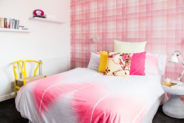 Bright, playful bedroom featuring a bed dressed in a pink-and-white patterned duvet and colourful decorative cushions. The room has a bold pink plaid feature wall, white shelving with books and objects, a yellow chair, and a white bedside table with a lamp.