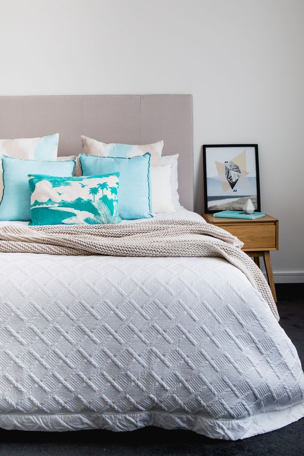 A bright, neatly styled bedroom featuring a large upholstered taupe headboard, layered white bedding, a chunky textured beige throw, and turquoise accent pillows. A simple timber bedside table holds framed artwork and small decor. The room has pale walls, dark carpet, and a clean contemporary coastal feel.