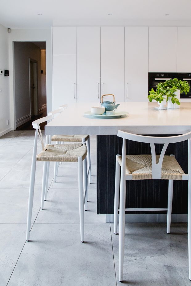 Bright, minimalist kitchen with a large pale island, integrated white cabinetry, black vertical-panelled island base, and white bar stools with woven seats. A built-in black oven and fresh green plants add contrast, while large light-grey floor tiles and recessed ceiling lights create a clean contemporary feel. An open doorway leads to a hallway.