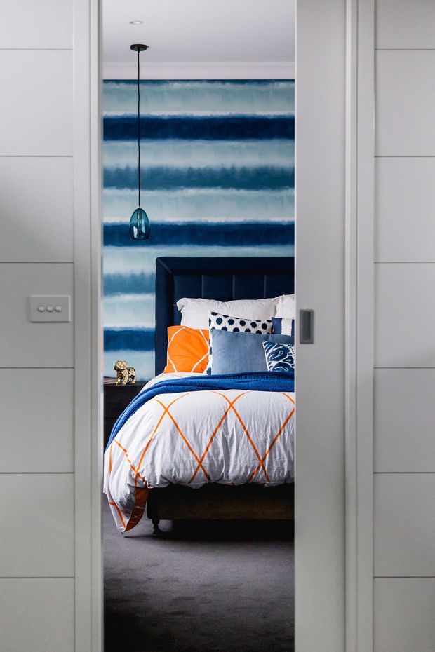 View through a partially open doorway into a stylish bedroom. The room features a dark navy upholstered bedhead, layered white and blue bedding with bright orange geometric accents, a blue-and-white abstract feature wall, a hanging glass pendant light, and grey carpet. White horizontal wall panelling and a sliding door frame surround the view.
