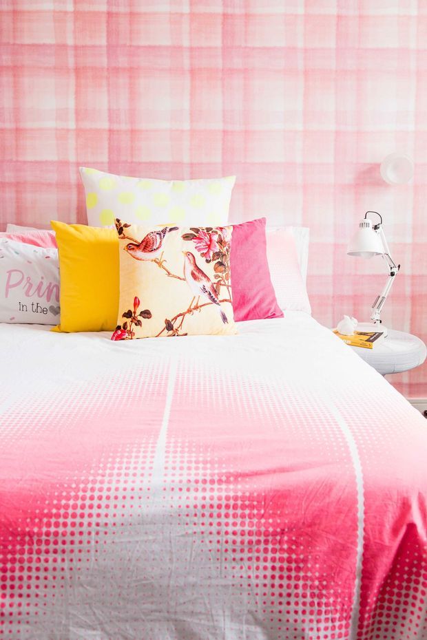 A brightly styled bedroom with a bed dressed in white and vivid pink bedding, surrounded by colourful patterned cushions including bird and floral motifs. The wall is covered in a soft pink plaid pattern, and a small round white bedside table holds a contemporary adjustable desk lamp and a book. The overall look is playful, youthful, and colourful.