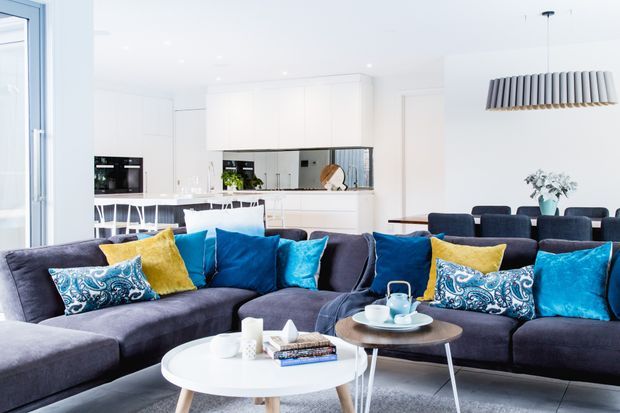 Bright open-plan living room with a large charcoal sectional sofa, colourful blue and yellow cushions, two round coffee tables, and a dining area beyond. A white contemporary kitchen with island seating, integrated appliances, mirrored splashback, and pendant lighting is visible in the background.