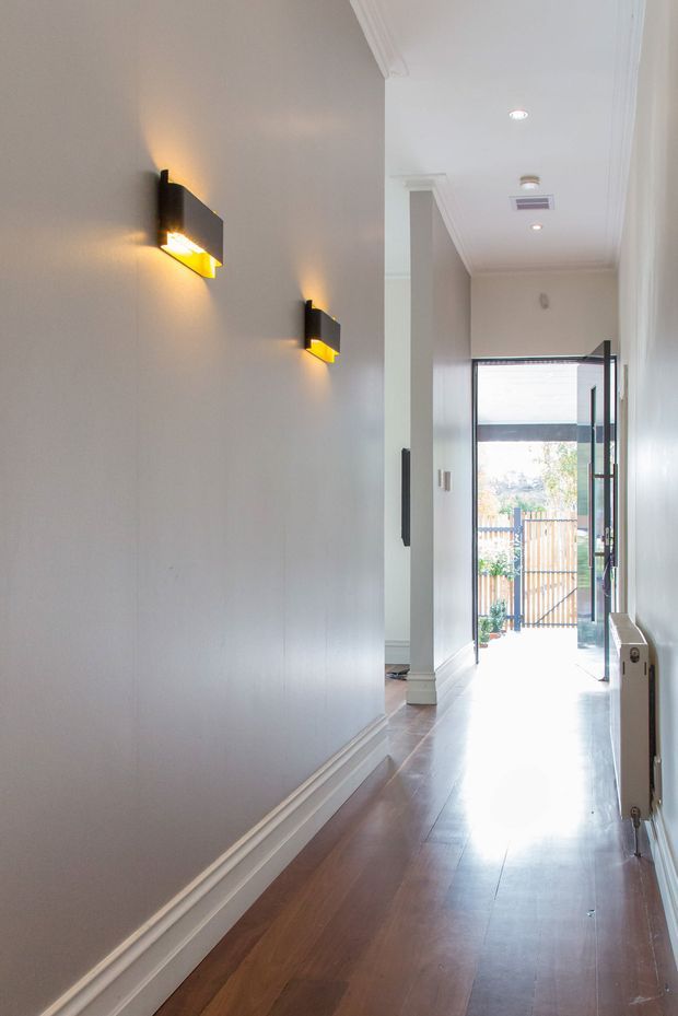A bright, narrow residential hallway with polished dark timber flooring, pale grey walls, white decorative skirting and ceiling cornices, and modern black-and-gold wall sconces. The corridor leads to an open glazed door and a small fenced outdoor area beyond.