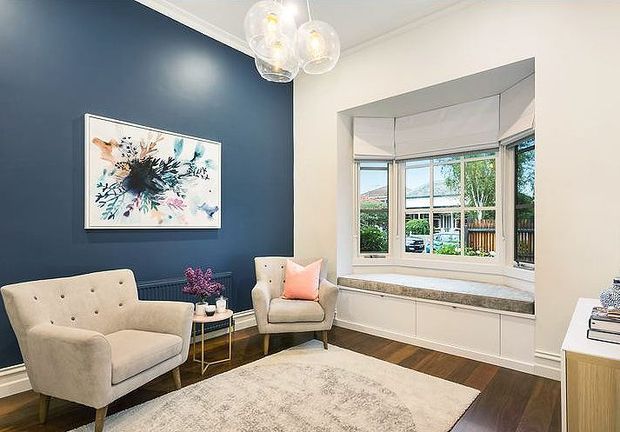 A refined living room with a deep navy feature wall, white decorative trim, pale upholstered armchairs, a small glass-and-metal side table, and a large light rug over dark timber flooring. A boxed bay window with integrated white cabinetry and a cushioned window seat brings in natural light, while clustered glass pendant lights and abstract artwork add decorative detail.