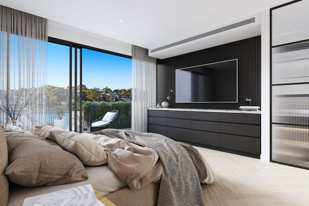 A contemporary bedroom with a low platform bed dressed in neutral beige bedding and textured throws, facing a wall-mounted television above dark built-in cabinetry. The room features full-height glazing with sheer curtains, a black vertical-slatted feature wall, pale timber flooring, recessed lighting, and a glazed black-framed wardrobe or partition. A leafy suburban landscape is visible outside.