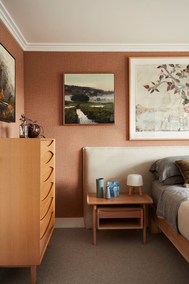 A warmly styled bedroom corner with a textured terracotta-brown wall covering, white decorative ceiling moulding, a large upholstered bedhead, and layered neutral bedding. A mid-century-inspired timber bedside table with open shelving and a small lamp sits beside the bed, while a timber chest of drawers and framed artwork complete the room. The space has a refined, cozy, art-focused character.