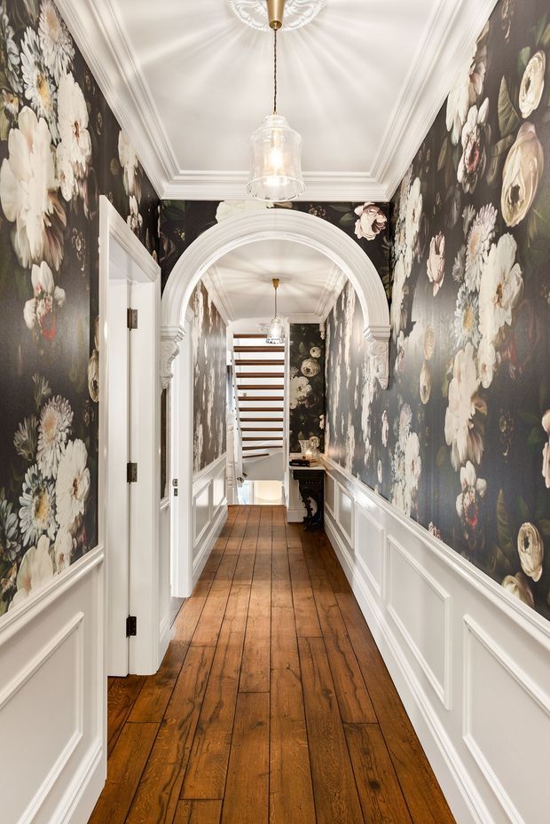 A narrow, richly decorated hallway with dark floral wallpaper, white raised-panel wainscoting and trim, warm timber plank flooring, ornate arched openings, and a coffered or tray ceiling with pendant lighting. The corridor leads toward a stairway and a small console area, creating a layered, traditional residential interior.