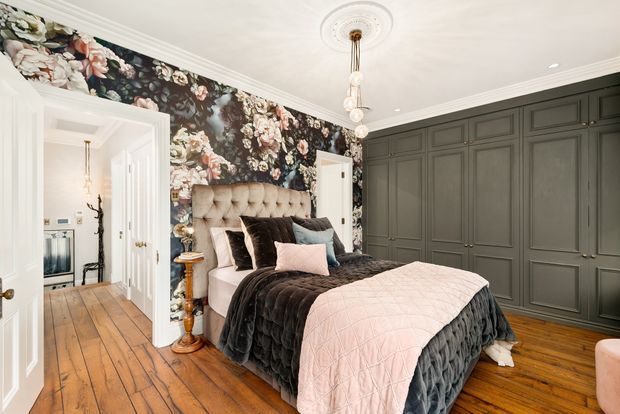 A refined bedroom with a tufted upholstered bed, layered dark and blush bedding, dramatic floral wallpaper, extensive dark built-in cabinetry, and warm timber floorboards. Decorative ceiling mouldings, a central pendant light, and white trim create a classic yet glamorous interior.