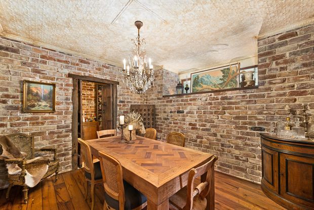 A richly detailed dining room with exposed red-brown brick walls, a patterned textured ceiling, and warm timber flooring. A large parquet-style timber dining table with woven and carved wooden chairs occupies the centre, illuminated by an ornate crystal chandelier. Decorative artwork, ceramics, candles, and a dark timber sideboard enhance the historic, rustic character.