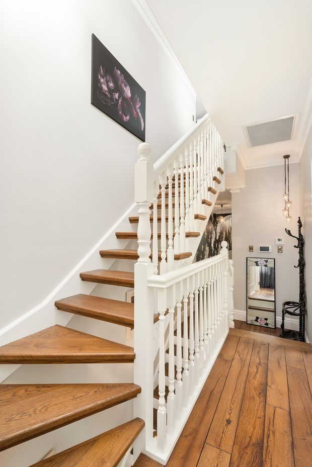 Bright residential stairway and landing with warm timber stair treads and flooring, ornate white painted balustrades and newel posts, white walls with decorative cornice, framed floral artwork, a mirror, and pendant lighting.