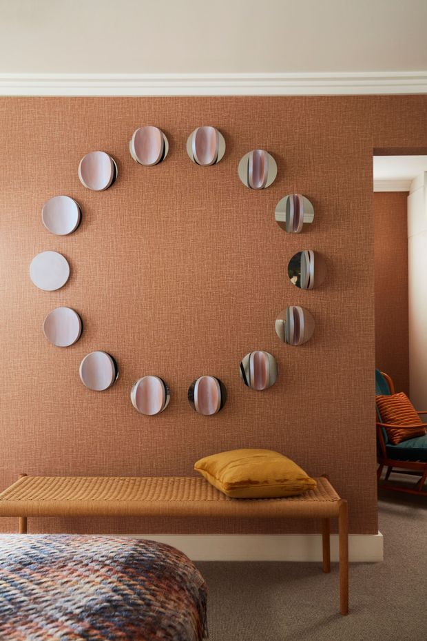 Warmly styled bedroom featuring a textured terracotta-brown feature wall with a circular arrangement of convex decorative mirrors. A woven timber bench with a mustard cushion sits beneath the wall, while the foreground shows a patterned bedspread. A doorway at right reveals part of an adjacent seating area with a teal chair and orange cushions. The room has traditional decorative ceiling mouldings and a cozy, richly layered palette.