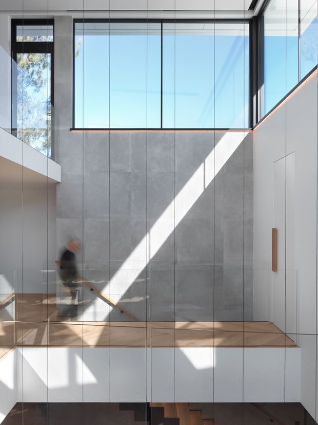 A bright, double-height contemporary stair hall with expansive black-framed glazing, pale grey vertically panelled walls, white built-in wall surfaces, and timber stair treads. Strong diagonal sunlight crosses the feature wall and stairs, while a blurred person moves along the stair landing. A lower-level stair is partially visible beneath the landing.