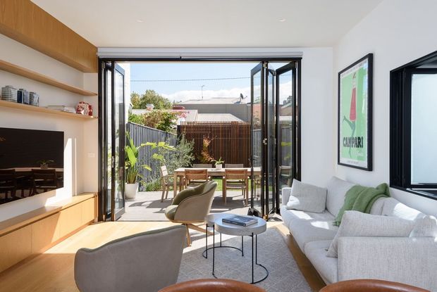 Bright contemporary living room with a built-in timber media wall and open shelving, a light upholstered sofa and armchairs, and black-framed folding glass doors opening onto a small outdoor dining terrace and courtyard. The room has white walls, a pale carpet, abundant natural light, and a framed graphic artwork.