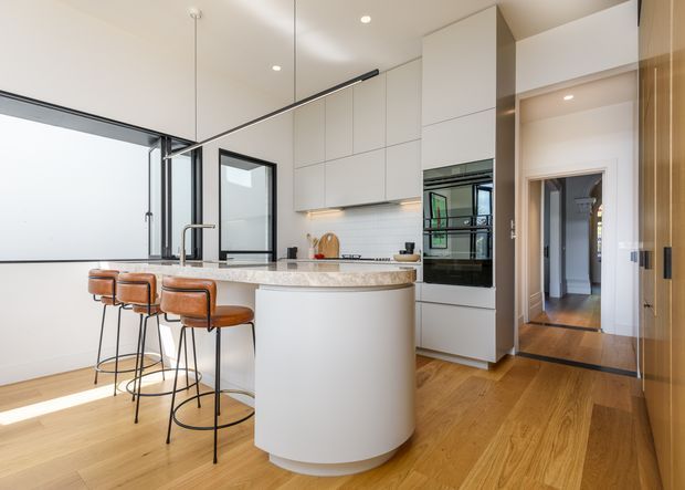A bright contemporary kitchen with a large curved island featuring a pale stone benchtop and integrated cabinetry, three brown leather bar stools, white handleless overhead cupboards, a tiled splashback, and built-in black appliances. Warm timber flooring continues into the hallway, while full-height glazing and black-framed internal windows add contrast. Recessed ceiling lights and a slim linear pendant provide illumination.