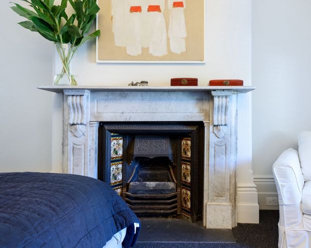 A traditional bedroom interior centered on an ornate white marble fireplace with a decorative tiled firebox and dark surround. A blue quilted bed occupies the foreground, with a white upholstered chair at right, a tall leafy plant in a glass vase, and framed abstract artwork above the mantel. The room has pale walls, white architectural trim, and dark carpeting.