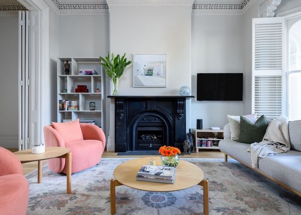 A bright, refined living room in a period-style home, featuring ornate ceiling cornices, a black decorative fireplace, built-in shelving, tall shuttered windows, and a contemporary seating arrangement. Pink armchairs, a grey sofa, a round timber coffee table, and a large patterned rug create a soft, eclectic interior.