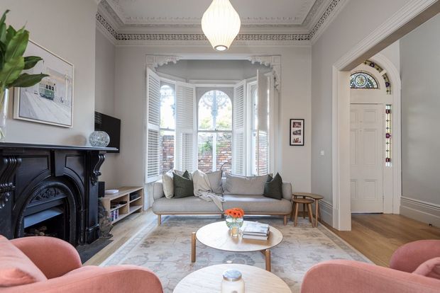 Elegant period living room with a black ornate fireplace, decorative ceiling cornice, pale neutral walls, timber flooring, a bay window with white shutters, and a light grey sofa arranged around a round coffee table. A detailed stained-glass panel surrounds the entry door, while blush pink armchairs and layered rugs add soft colour and comfort.