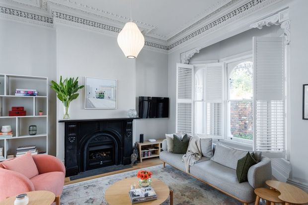 Bright, elegantly detailed living room with ornate Victorian-style ceiling cornices and decorative window architraves, a black fireplace, white plantation shutters, built-in shelving, a grey sofa, pink armchair, and a round timber coffee table. The room has a refined yet inviting character with abundant natural light.