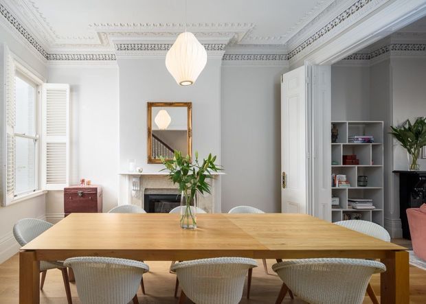 Bright, elegant dining room with a long natural timber dining table surrounded by woven upholstered chairs. The room features ornate white decorative ceiling cornices and mouldings, a classic fireplace with marble surround, large shuttered windows, built-in shelving, a pendant light, and a gold-framed mirror. The styling blends refined traditional architecture with contemporary furnishings and warm natural accents.