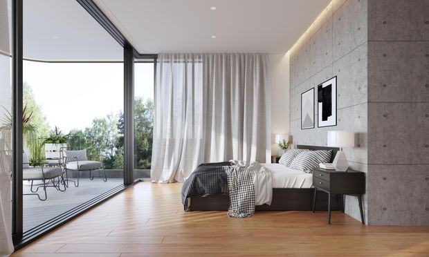 A contemporary bedroom with a low-profile bed, dark bedside table, soft drapery, and minimalist artwork. Full-height sliding glass doors open to a terrace, bringing in abundant natural light. The room features warm timber flooring, a raw concrete feature wall, recessed ceiling lighting, and a neutral black-and-white palette.