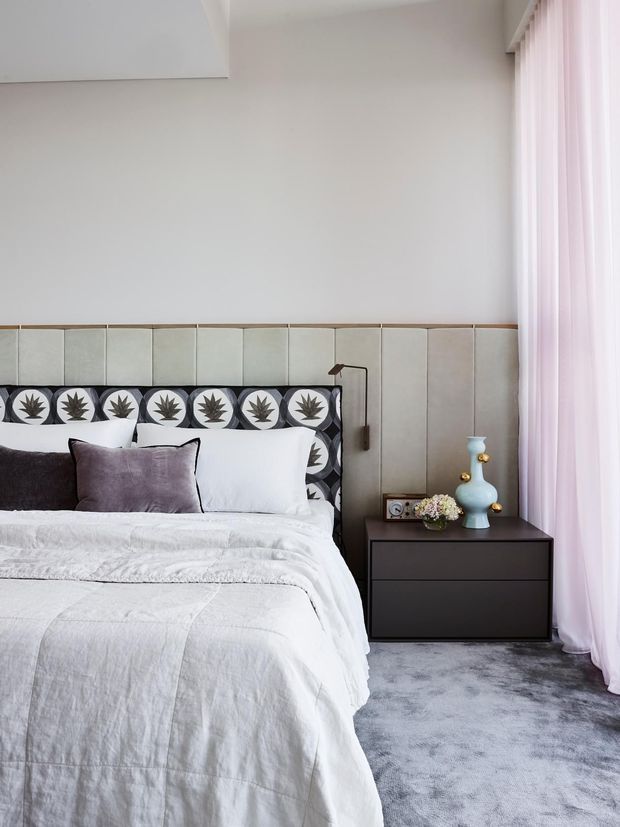 A refined contemporary bedroom with a large upholstered and patterned headboard, white layered bedding, dark bedside cabinetry, a sculptural blue table lamp, and full-height pale pink curtains. The room has a soft neutral palette with grey carpeting and subtle decorative detailing.