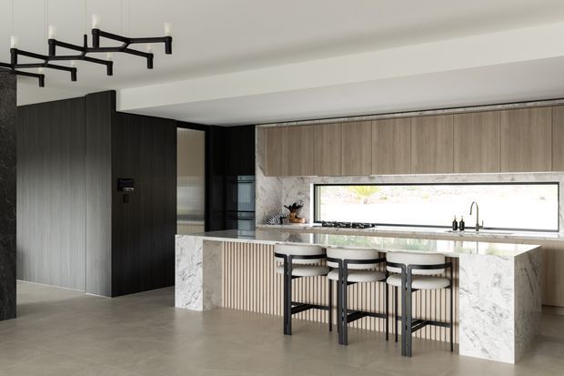 A contemporary open-plan kitchen featuring a large marble-look island with waterfall ends, vertical fluted detailing, dark timber cabinetry, light timber overhead cabinets, black-framed glazing, and three bar stools. A sculptural black linear pendant hangs above the island, while a long horizontal window provides natural light over the benchtop and sink.
