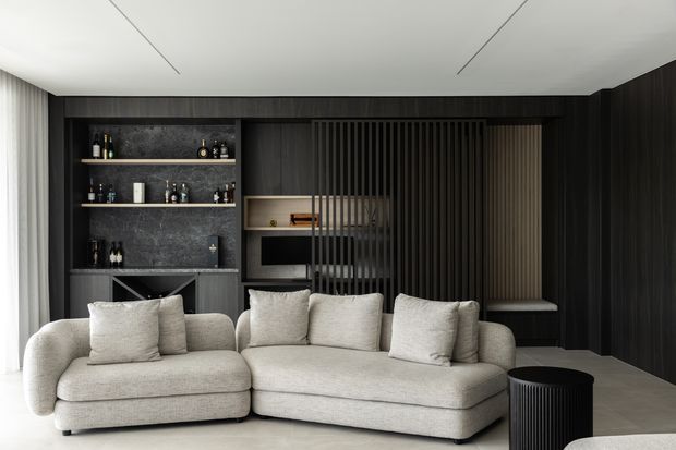A refined contemporary living room with a large cream sectional sofa facing a dark built-in media wall. The wall combines vertically ribbed dark timber panelling, open display shelving with a dark stone-like backing, a concealed television niche, and a slatted screen partially dividing the space. Pale curtains, light flooring, and a clean white ceiling balance the dark joinery. A small black ribbed side table sits beside the sofa.