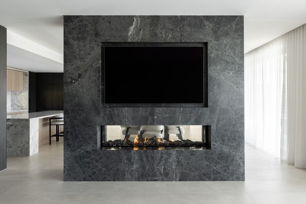 Contemporary open-plan living area centred on a dark grey marble fireplace and media wall. A linear gas fire is built into the stone feature wall beneath a recessed television, with a bright dining or living space to the right and a partial kitchen with island seating visible to the left. Floor-to-ceiling sheer curtains and pale flooring create a refined, high-contrast interior.