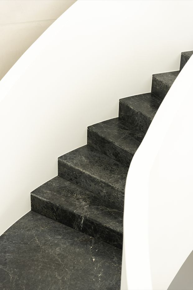 Minimalist interior stairway with dark veined stone steps and matching stone risers, enclosed by smooth curved white walls and a sculptural white balustrade. The composition emphasizes the geometric sweep of the staircase and strong contrast between the dark marble-like stone and bright surfaces.