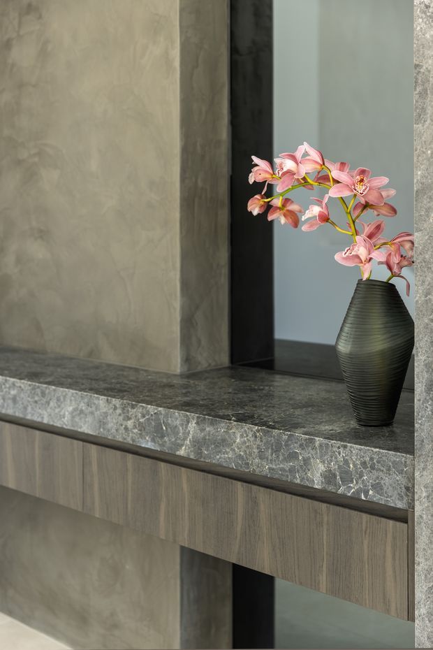Close-up of a refined built-in console or vanity with vertically grained wood cabinetry, a dark grey veined stone benchtop, textured grey wall surfaces, and a large black-framed mirror. A ribbed black vase with pink orchid branches provides a decorative accent. The composition is minimalist and moody, emphasizing layered stone, timber, and reflective surfaces.