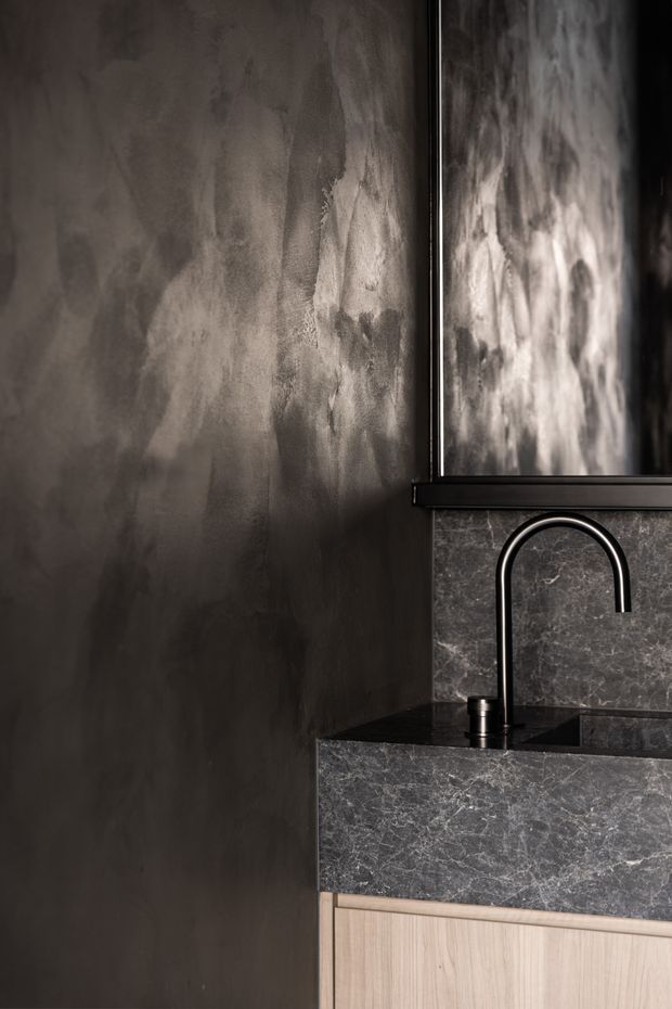 Close-up of a moody, refined bathroom vanity featuring a dark stone basin and backsplash, black arched tapware, light timber cabinetry, a textured dark plaster wall, and a large framed mirror.