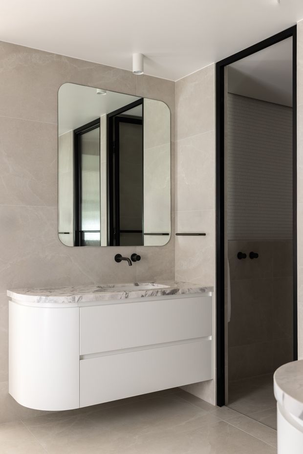 Contemporary bathroom with a floating white curved-front vanity, veined marble benchtop, large rounded-corner mirror, black wall-mounted tapware and hardware, and a dark-framed shower enclosure. Large warm grey-beige stone-look tiles cover the walls and floor, creating a refined, understated interior.