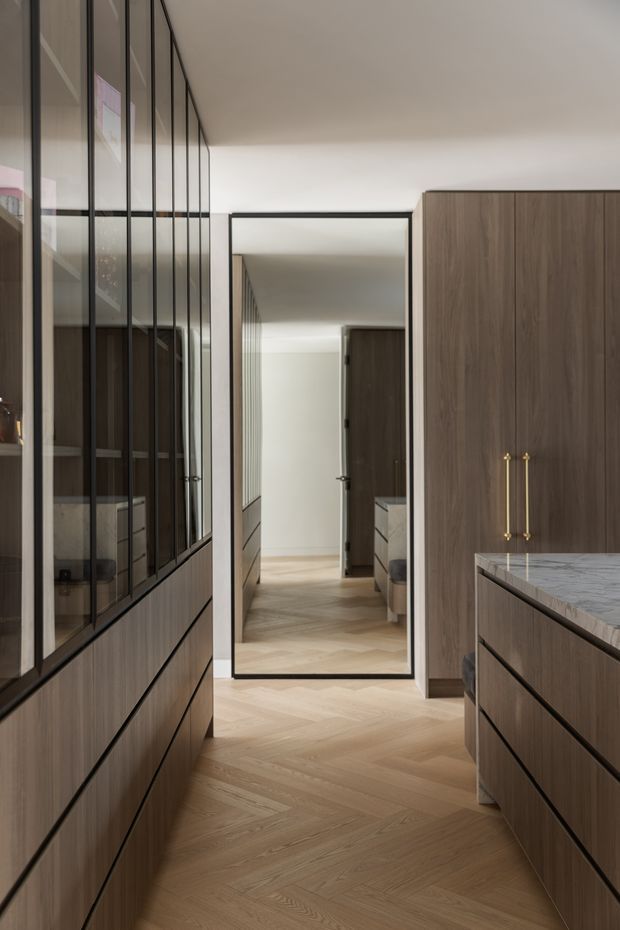 A narrow, refined kitchen or kitchen passage with dark timber cabinetry on both sides, integrated black-framed glass-front storage along the left wall, a veined light stone benchtop on the right, and warm herringbone timber flooring. A full-height black-framed mirror or glazed opening at the end visually extends the space. The palette is warm and neutral, with minimalist detailing and brass cabinet pulls.