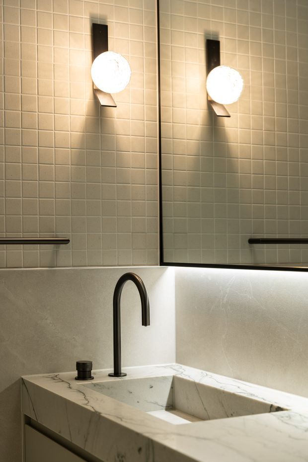 Close-up view of a refined bathroom vanity with a sculptural white marble countertop and integrated rectangular basin. A tall matte-black curved faucet and matching small fixture contrast with the pale stone. The wall is clad in small square beige tiles, with a large mirror, warm globe wall sconces, and a dark horizontal towel rail. Under-vanity lighting creates a warm, softly lit atmosphere.