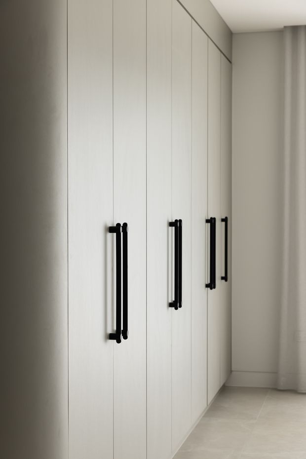A minimalist hallway or dressing area lined with tall, floor-to-ceiling white built-in cabinets. The cabinetry has slim vertical panel divisions and contrasting black elongated pull handles. The space features pale neutral walls, a smooth white ceiling, light tiled flooring, and a softly lit, clean contemporary appearance.