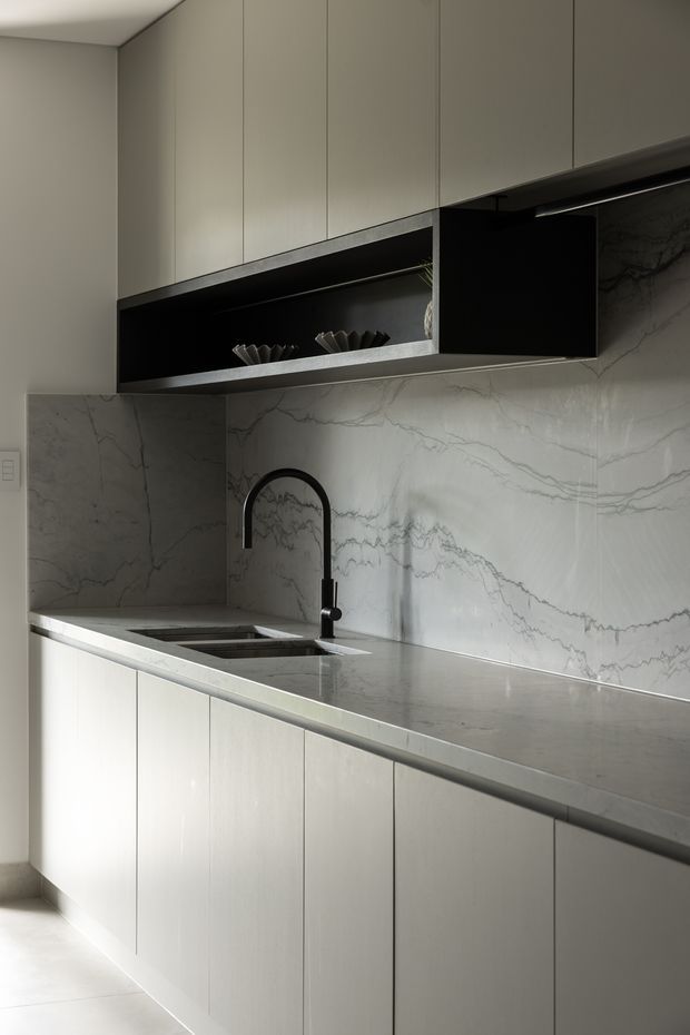 A refined, minimalist kitchen with full-height matte grey cabinetry, integrated open display shelving, a long grey-veined stone benchtop and matching full-height splashback. A black gooseneck tap and undermount sink provide contrast, while concealed lighting and clean handleless joinery create a streamlined appearance.
