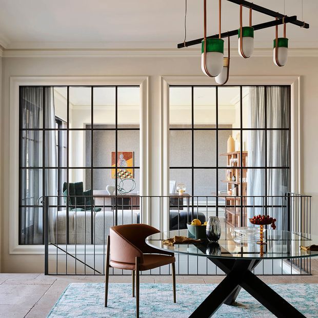 A refined dining area with a glass-top dining table and sculptural black X-shaped base, surrounded by upholstered brown dining chairs. Large black-framed glass partitions divide the dining space from a living room, with layered curtains, a pale patterned rug, built-in wall detailing, and a distinctive linear pendant light with green and white shades. The room combines classic architectural mouldings with contemporary furnishings and metal-framed glazing.
