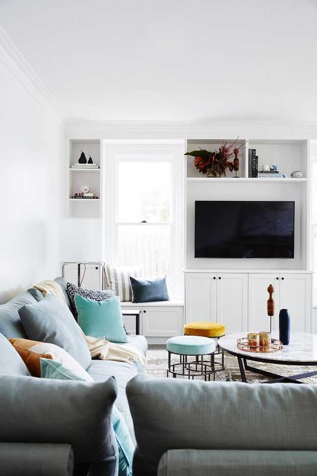 Bright, refined living room with extensive white built-in cabinetry surrounding a television and window seat. The room features a large pale upholstered sectional sofa, layered blue and teal cushions, a round coffee table, pastel upholstered stools, and carefully styled shelving with decorative objects and books. Traditional moulded wall and ceiling details are paired with a fresh contemporary interior palette.