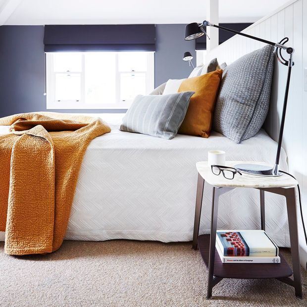 Bright, well-styled bedroom with a neatly made bed, layered grey and mustard cushions, and a textured mustard throw. The room has dark blue-grey walls, white window trim and bedding, beige carpet, and a slim two-tier bedside table with an adjustable black reading lamp and books.