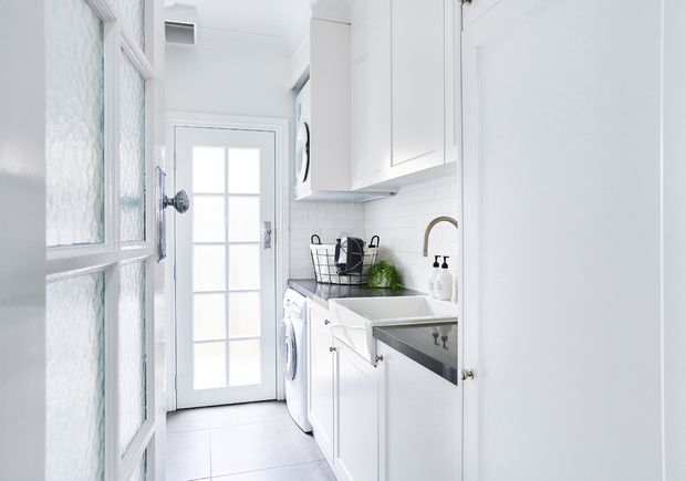 Bright, compact white laundry room with built-in cabinetry, a dark countertop, integrated laundry appliances, a deep utility sink, subway-tile backsplash, and a glazed exterior door. The space has a clean, traditional-style fit-out with abundant natural light.