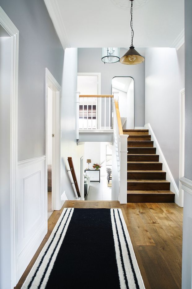 Bright, narrow residential hallway and stairway with pale grey walls, white decorative wainscoting and trim, warm timber flooring, and a dark wood staircase with white balustrades. A long black-and-white runner leads toward the stairs, while pendant lights and a tall arched mirror add visual interest. The hall opens to a lower living area and an upper landing.