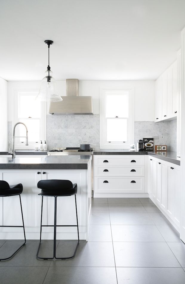 Bright, compact contemporary kitchen with white shaker-style cabinetry, a dark stone island benchtop, pale marble-look tiled splashback, large light-grey floor tiles, stainless-steel rangehood, and black bar stools. A glass pendant light hangs above the island, with abundant natural light from multiple windows.