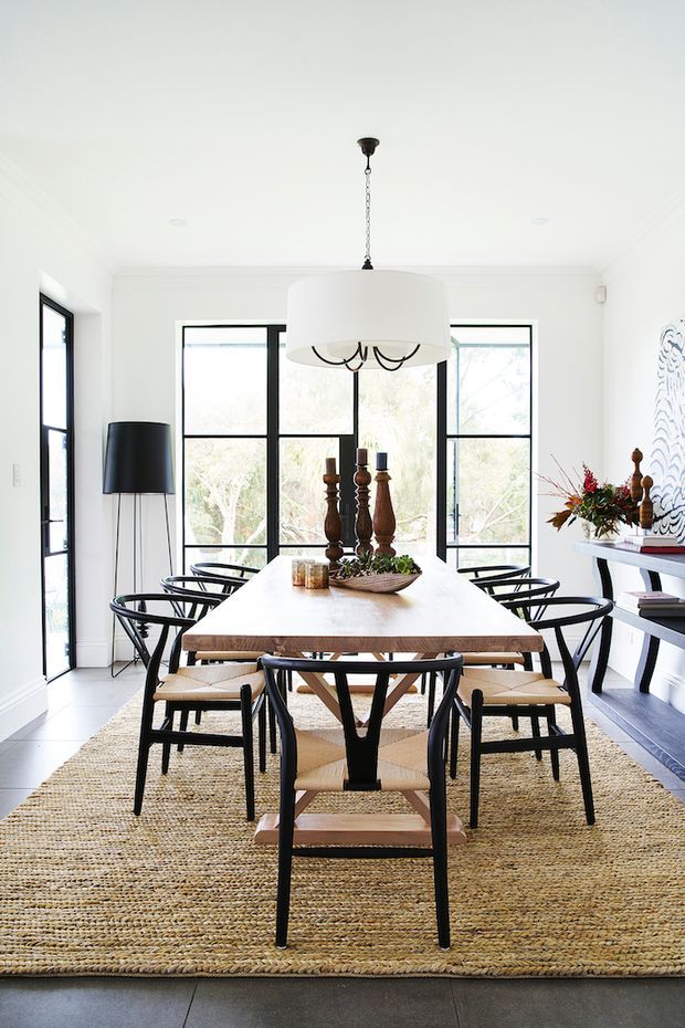 Bright, refined dining room with a long light timber dining table surrounded by black-framed woven chairs. Large black-framed windows and a glazed door provide abundant natural light, while a large white pendant, woven rug, dark floor lamp, built-in shelving, and decorative centerpiece complete the room.